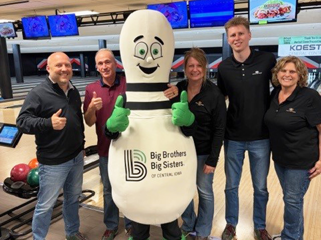 A group of adults in a bowling ally standing by a bowling pin mascot.